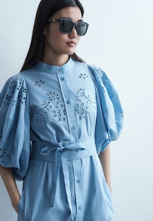 Žena nosi čierne slnečné okuliare a modré šaty s kvetinovou výšivkou a opaskom zviazaným v páse, otočená doľava.