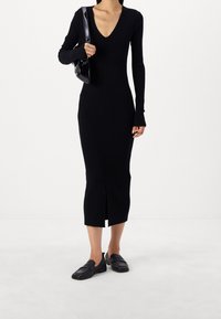 Svart ribbade midi-klänning med långa ärmar, djup V-ringning och sprund framtill; matchad med svarta slip-on skor och en liten handväska.