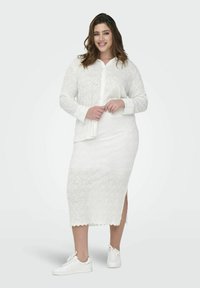 Witte jurk met een ingewikkeld geborduurd rozenpatroon, lange mouwen, knoopsluiting aan de voorkant en zijsplitten. Gecombineerd met witte sneakers.