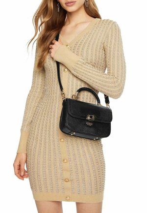 Femme portant une robe en maille beige texturée avec boutons et portant un sac à main en cuir noir avec une quincaillerie dorée sur l'épaule.