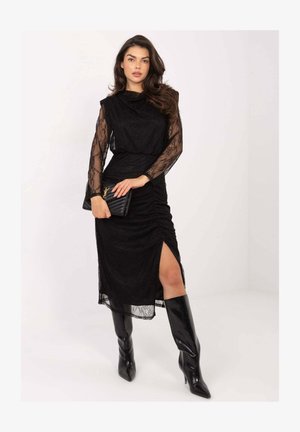 Schwarzes Spitzenkleid mit langen, transparenten Ärmeln, einem gerafften Taillenbereich und einem seitlichen Schlitz. Kombiniert mit schwarzen knielangen spitzen Stiefeln und einer Clutch.