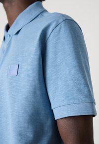 Polo de cor azul clara feito de tecido texturizado, apresentando um emblema em relevo no peito e mangas curtas com punhos canelados.