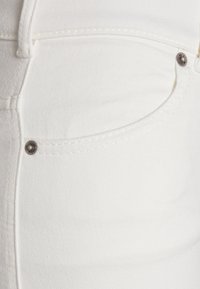 Vit denimtyg av en byxa, med en framficka med metallnitar och sömdetaljer, som visar en slät textur och ren design.