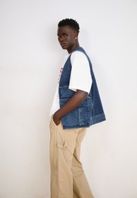 Denim brezrokavnik z modrim pranjem, ki se ponaša z udobnim krojem in odprtim sprednjim delom; v kombinaciji z belim majico in bež vojaškimi hlačami.