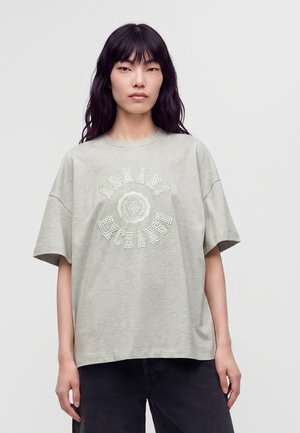 Jeune femme aux longs cheveux foncés porte un t-shirt ample gris clair Armani Exchange et un pantalon foncé sur un fond uni.