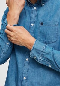 Muž upravující rukáv modré džínové košile s dlouhým rukávem, zaměřující se na detaily rukou a košile.