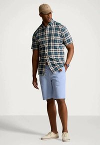 Short-sleeve plaid shirt in green, navy, and beige, paired with light blue shorts. Accessories include a beige cap and white sneakers.