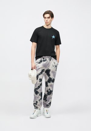 Melna kokvilnas T-krekls ar zilu zvaigžņu logotipu; pelēkas un melnas krāsas svītrainas sporta bikses; baltas kurpes; rokās tur bēša cepuri.