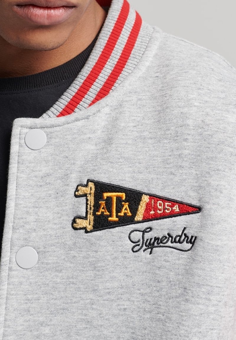 Gros plan d'une personne portant une veste grise avec un col à rayures rouges et un écusson brodé indiquant "ATA 1954 Superdry" sur la poitrine.
