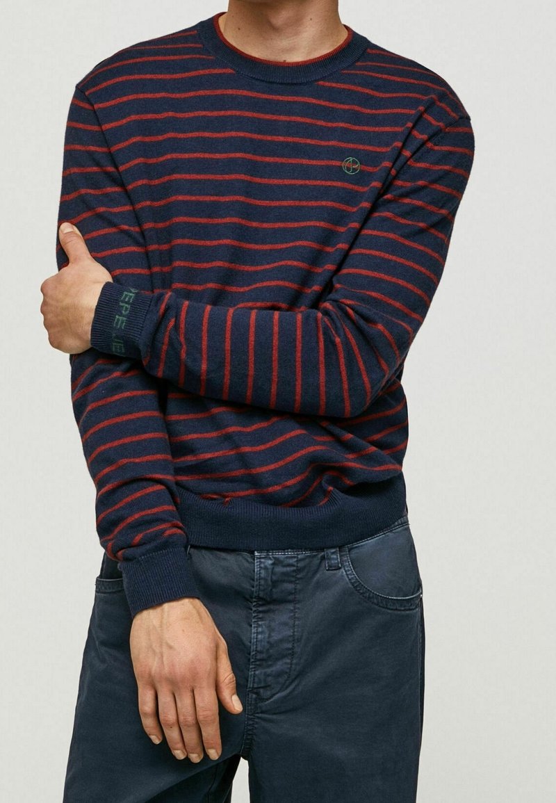 Navyblauer gestreifter Pullover mit roten horizontalen Linien, gerippten Bündchen und Saum, mit einem kleinen grünen Logo auf der Brust. Das Material wirkt weich.