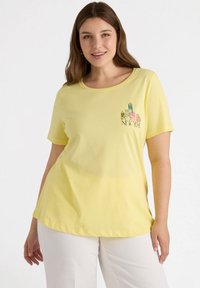 Camiseta de algodón amarilla con mangas cortas y cuello redondeado, que presenta un gráfico bordado de "New York" en detalles multicolores en el pecho.