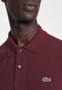 Burgundy színű poloing, texturált anyagból, két gombos nyakkal és egy kis hímzett krokodil logóval a mellrészen.
