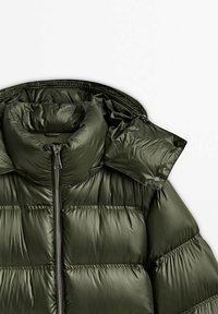 Vihreä puffer-takki, jossa on korkea kaulus, vetskareita ja neppareita. Sileä, kiiltävä kangas, jossa on tikkauksia ja rakenteellinen huppu.