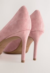 Paar roze suède hoge hakken met puntige neuzen en slanke hakken, van achteren getoond op een lichte achtergrond.