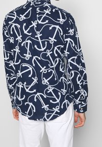 Chemise navy avec un motif d'ancre blanche et de corde, manches longues, poignets à boutons et ourlet incurvé. Fabriquée en tissu léger.