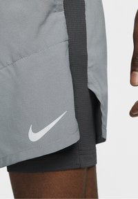 Shorts gris clair avec un swoosh Nike blanc superposé sur des shorts noirs sur la partie supérieure de la cuisse d'une personne et main sur le côté.