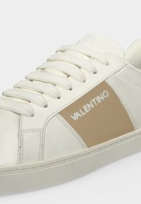 Baskets blanches avec panneau latéral beige et lacets blancs, avec le nom de la marque "Valentino" sur le côté.