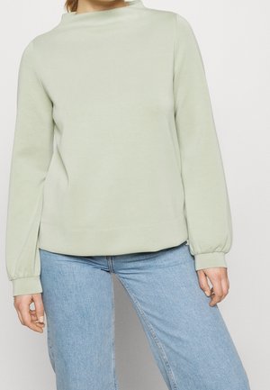 Kvinde iført en lysegrøn langærmet sweatshirt med høj hals og lyseblå jeans mod en ensfarvet baggrund.