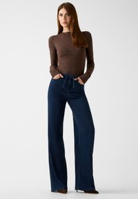 Bruine nauwsluitende top met lange mouwen en subtiel patroon, gecombineerd met een high-waist, wijdgespreide donkerblauwe denimbroek met een trekkoord.