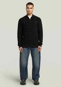 Pull noir à col zippé avec texture côtelée et manches longues, associé à un jean large bleu clair et des baskets noires avec des accents blancs.