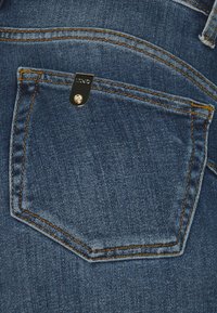 Bolsa trasera de unos vaqueros de mezclilla en azul oscuro con costuras naranjas, que presenta una etiqueta rectangular negra con herrajes metálicos y un botón plateado.