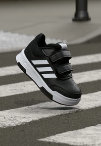 Scarpa atletica nera con superficie testurizzata, dotata di tre strisce bianche, cinturini regolabili in velcro e suola bianca.