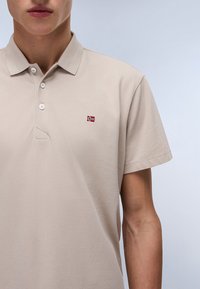 Světle béžové polo tričko vyrobené z texturované tkaniny. Obsahuje tříknoflíkovou légu a malou nášivku norské vlajky na levé části hrudi.