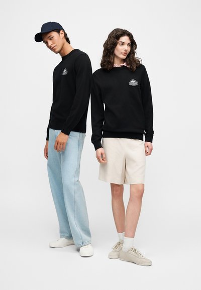 Un homme et une femme portant des sweatshirts noirs Lacoste, l'homme en jean bleu clair et casquette, la femme en short beige et baskets, debout côte à côte.