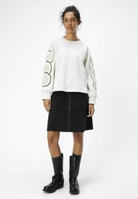 Witte sweatshirt met patroon op de mouwen, gecombineerd met een zwarte denim rok. Zwarte kniehoge laarzen maken de outfit compleet.