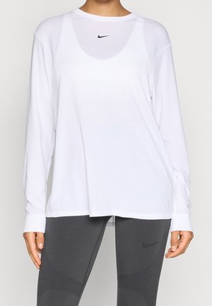 Osoba ubrana w białą, długorękawową koszulkę Nike oraz ciemnoszare legginsy Nike, stojąca na jednolitym tle.