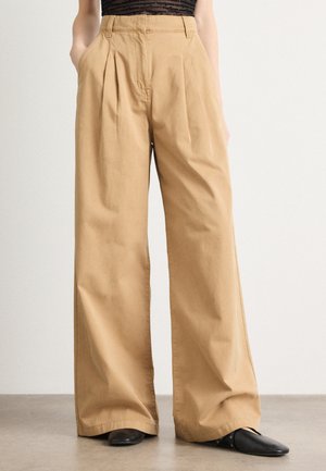 Bestrækkede bukser i tan lavet af bomuld, med for-placerede folder, sidelommer og høj talje, parret med sorte spidse sko.