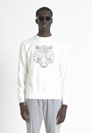 Sudadera blanca con un gráfico de tigre negro detallado en la parte frontal. Combinada con pantalones grises claros con pliegues y gafas de sol oscuras.
