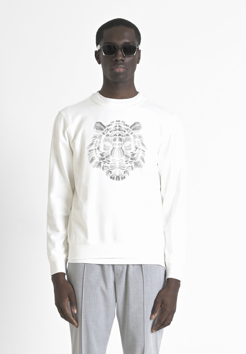 Sudadera blanca con un gráfico de tigre negro detallado en la parte frontal. Combinada con pantalones grises claros con pliegues y gafas de sol oscuras.