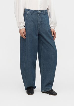 Person iført løse vidde-blå jeans, hvid langærmet bluse og sorte sko, der står mod en ensfarvet baggrund.