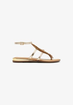 Guldmetalliske flade sandaler med flere smalle remme, der har en kvastdetalje og en brun sål med guldfarvet metaldetalje på hælen.