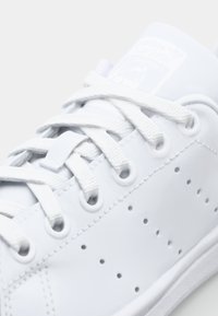 Bílé tenisky Adidas Stan Smith zblízka, ukazující tkaničky, perforované strany a logo na jazyku na bílém pozadí.