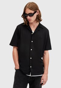 Black short-sleeve shirt with a relaxed fit, featuring white buttons and a subtle logo detail on the chest. Worn with black sunglasses.