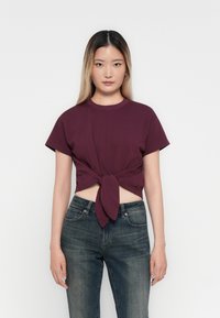 Bordeaux crop top met korte mouwen en een knoopdetail aan de voorkant, gecombineerd met donkerblauwe jeans, op een plain witte achtergrond. Glad stoftextuur.