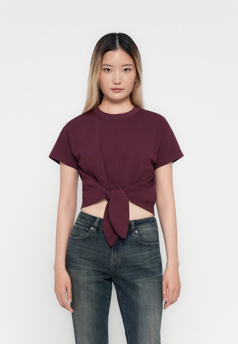 Bordeaux crop top met korte mouwen en een knoopdetail aan de voorkant, gecombineerd met donkerblauwe jeans, op een plain witte achtergrond. Glad stoftextuur.