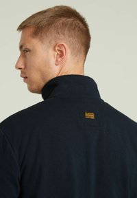 Sweatshirt colla blu navy con tessuto texturizzato. Presenta un piccolo tag di marca giallo e nero rettangolare sul retro.