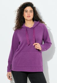 Donna con capelli ricci che indossa una felpa viola con lacci decorativi e pantaloni neri, in piedi su uno sfondo bianco.