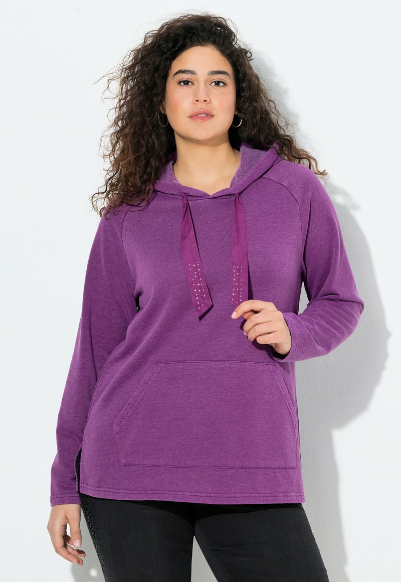 Donna con capelli ricci che indossa una felpa viola con lacci decorativi e pantaloni neri, in piedi su uno sfondo bianco.