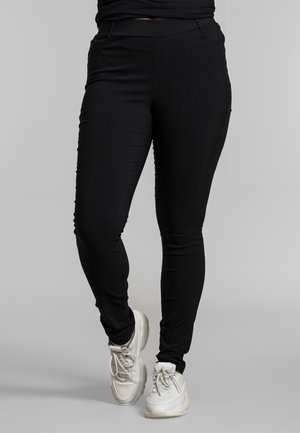 Osoba nosząca obcisłe czarne spodnie i białe masywne trampki na jednolitym szarym tle, stojąca z jedną lekko zgiętą nogą.