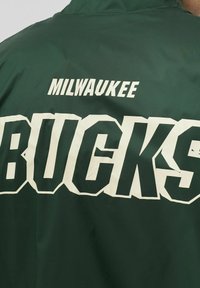 Giacca sportiva verde con grande scritta bianca "BUCKS" e "MILWAUKEE" sopra. Realizzata in tessuto liscio con finitura semi-matte.