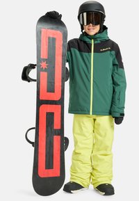 Snowboard s čiernym povrchom s červeným logom. Jazdec má na sebe zeleno-čiernu bundu, žlté nohavice a čierne rukavice.