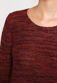 Pull en maille de couleur rouge foncé avec un motif texturé. Doté d'un col rond et de coutures visibles, créant un effet superposé.