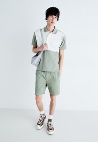 Zelené polo tričko s bielou panelou, skombinované so zelenými šortkami. Model má na sebe hnedé tenisky a drží malú šedú crossbody tašku.