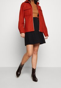 Chaqueta roja con cremallera y dos bolsillos frontales, llevada sobre un top de punto marrón y una falda negra con pliegues, combinada con botas de tobillo marrones y brillantes.