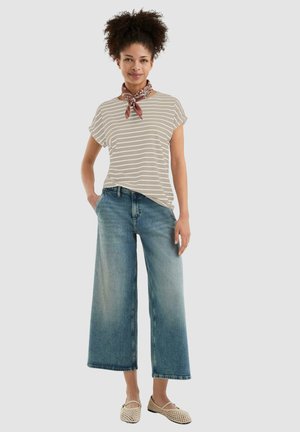 Gestreepte beige T-shirt, ruime pasvorm, gecombineerd met wijde blauwe denim jeans. Model draagt een rode patroon sjaal en lichtgekleurde schoenen.