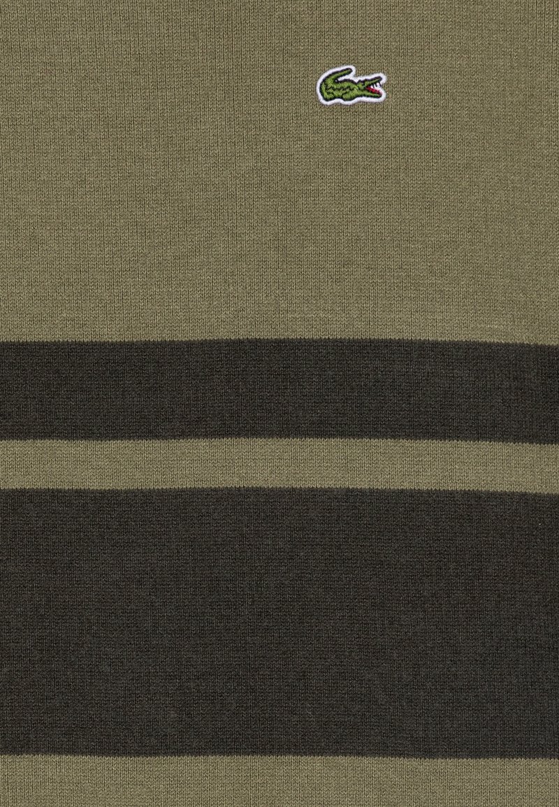 Pull vert olive foncé avec des rayures horizontales noires. Présente un petit logo de crocodile brodé en vert et rouge. Tissu tricoté doux et texturé.